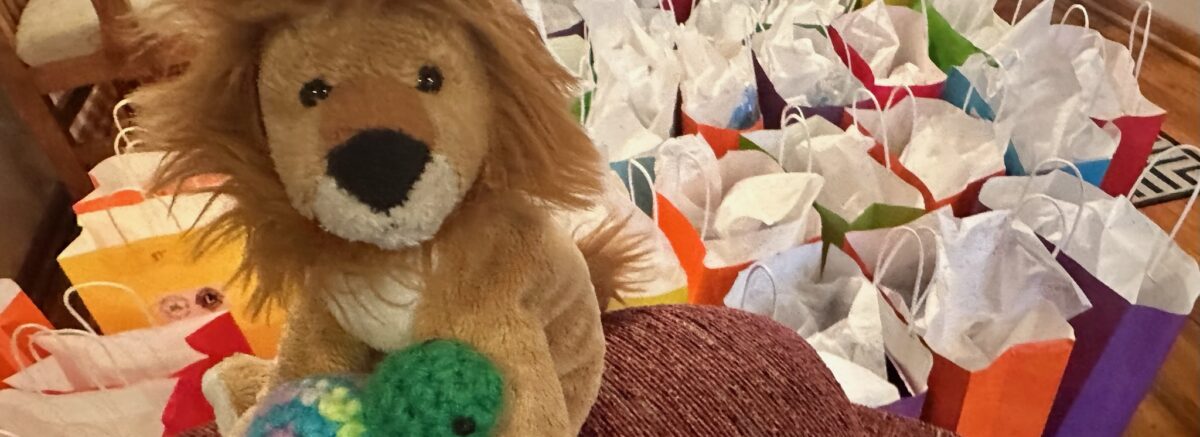 A toy lion holds a toy turtle with a number of gift bags on the floor in the background