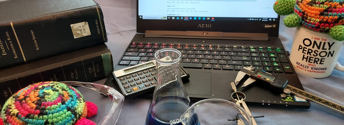 Two toy turtles, one sitting atop a coffee mug looking at a laptop screen, the other peering through oversized safety glasses at a flask and beaker filled with liquid, with textbooks and scientific instruments strewn about